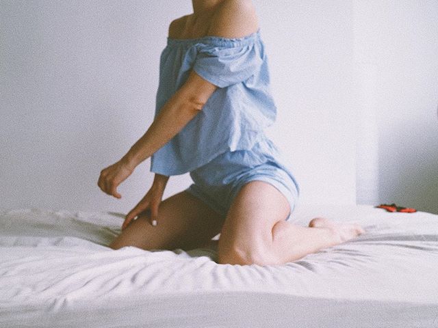 A woman in a blue outfit kneeling on a white bed in a minimalist setting.