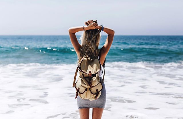 A woman with a backpack stands on the beach, looking out at the ocean.