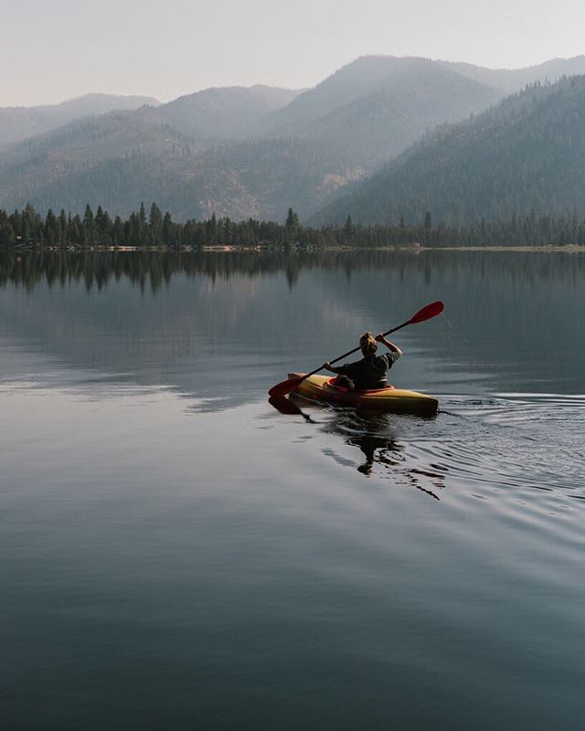 A lone kayaker paddles through a serene lake surrounded by misty mountains and lush forest.