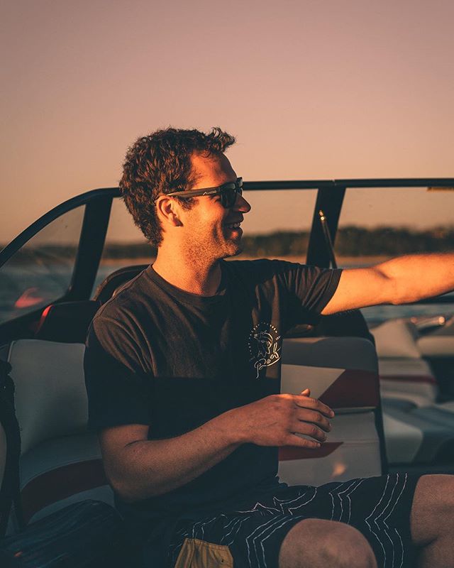 A smiling man enjoys a boat ride at sunset wearing sunglasses, relaxed and happy.