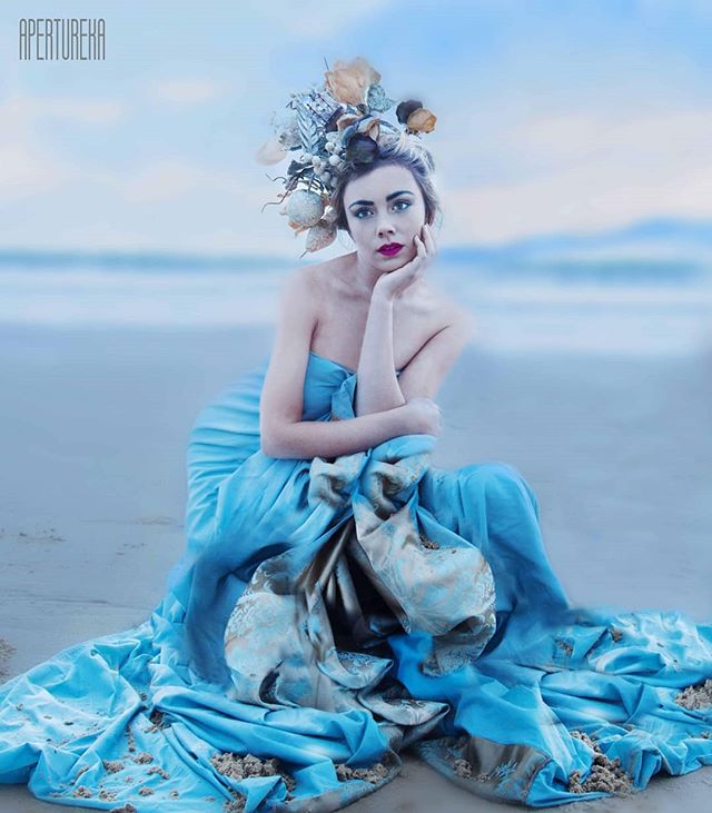 A woman in a blue dress and headpiece sits gracefully on a beach, creating a dreamy editorial image.
