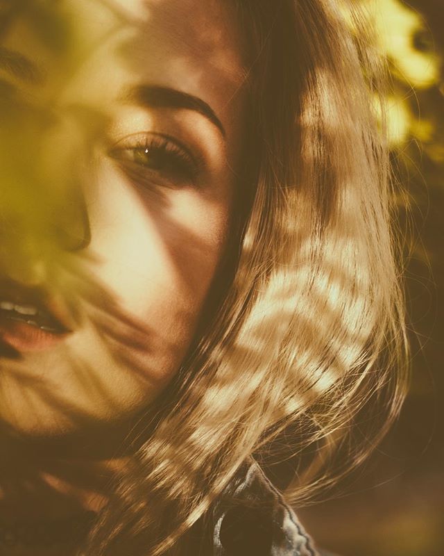 Close-up portrait of a woman with blonde hair, partially obscured by shadows and leaves, bathed in soft, warm light.
