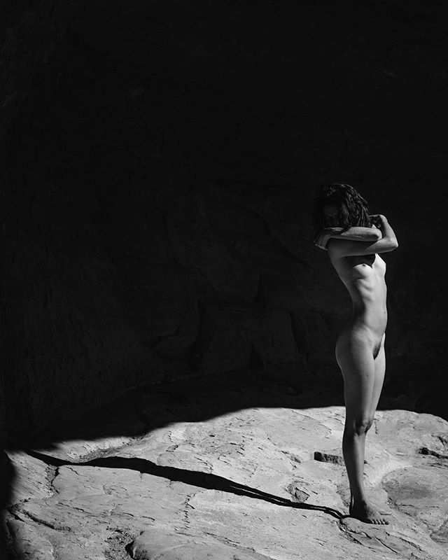 A nude woman stands on a rock in a fine art black and white image, bathed in soft, natural side lighting.