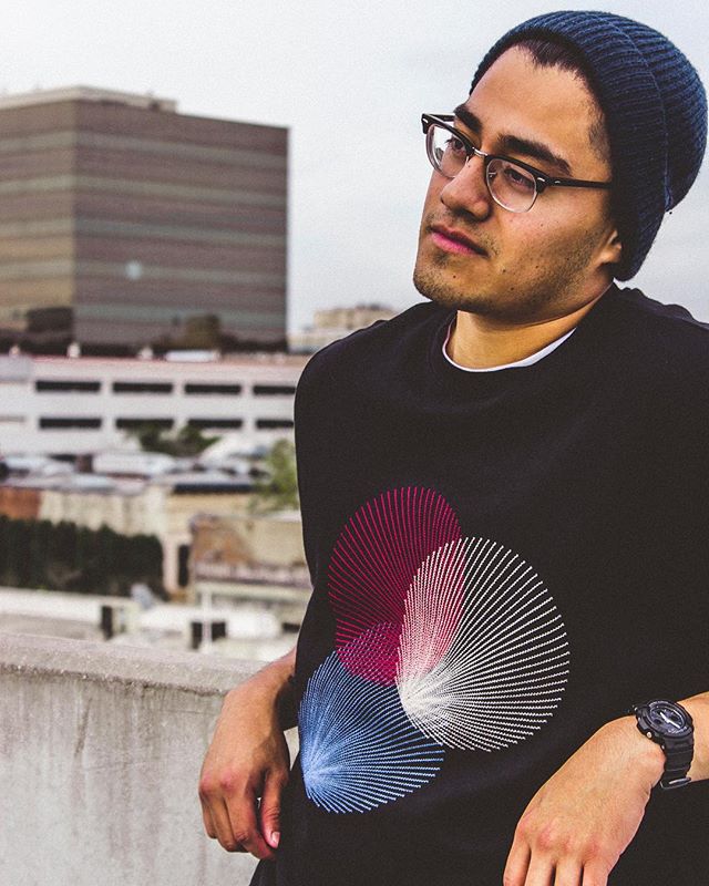 A man wearing glasses and a beanie stands on a rooftop with a cityscape backdrop.