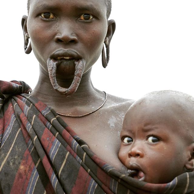 A woman with a lip plate holds her baby in a traditional setting, showing motherhood and cultural heritage.