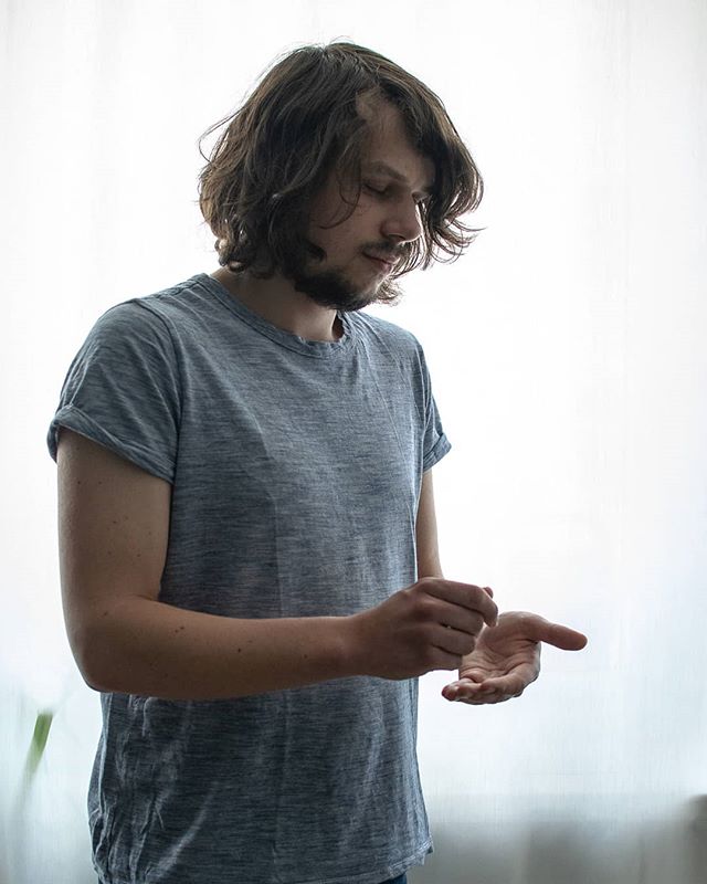 A man with long hair looks down at his hands in a soft, natural light setting.