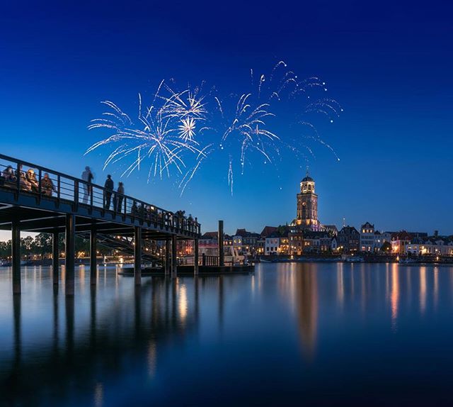 People watch fireworks over a city skyline at night, reflected in the water.