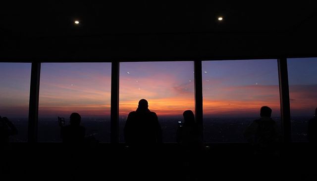 Silhouettes of people looking out at a vibrant sunset over a cityscape from a high vantage point.