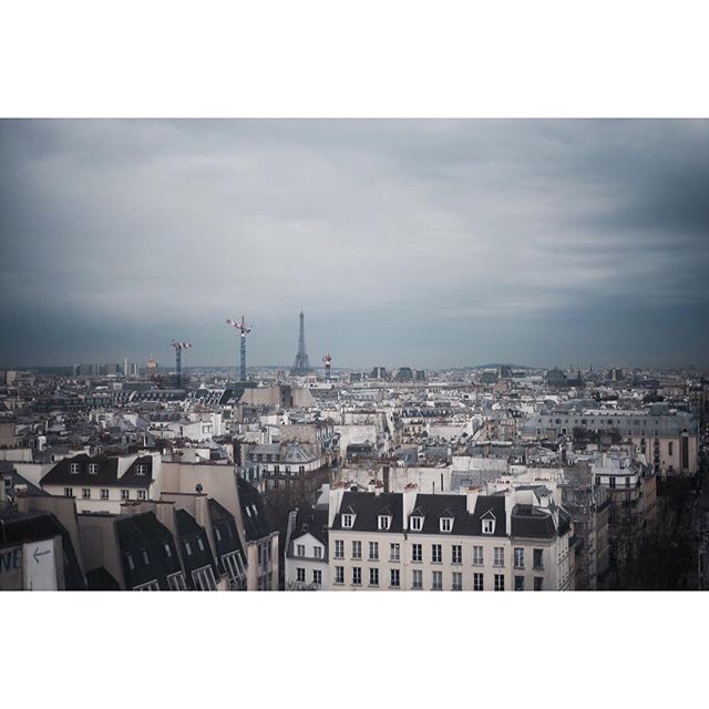 An aerial view captures the Paris cityscape under an overcast sky, featuring the Eiffel Tower and building rooftops.