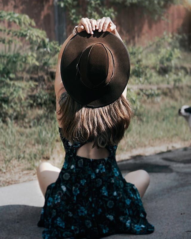 A woman in a floral dress sits outside holding a hat above her head in a relaxed pose.