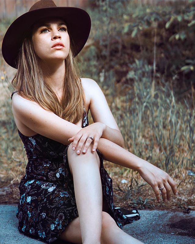 A woman in a floral dress and hat sits outdoors in soft light, looking up.
