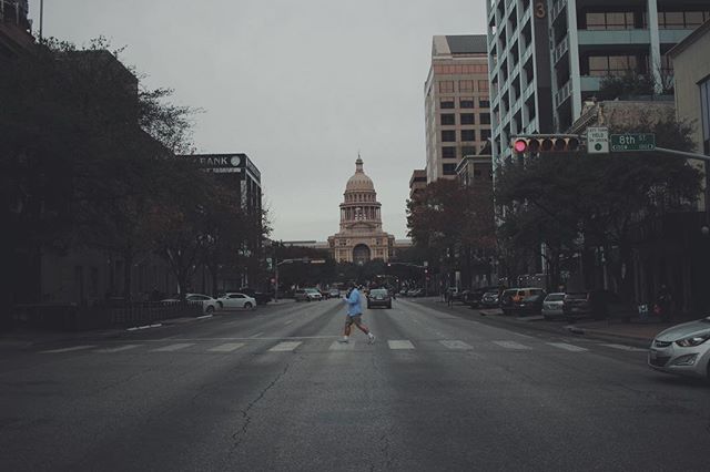 A man crosses a city street with the capitol building in the background on a cloudy day.