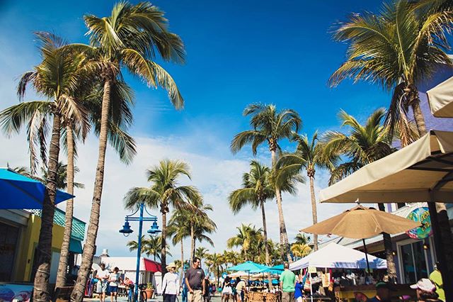 Vibrant tropical scene with palm trees, people, and umbrellas in a bustling beach town marketplace on a sunny day.