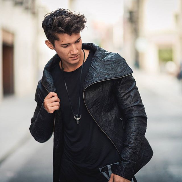 A young man in a black jacket and necklace poses on a city street.