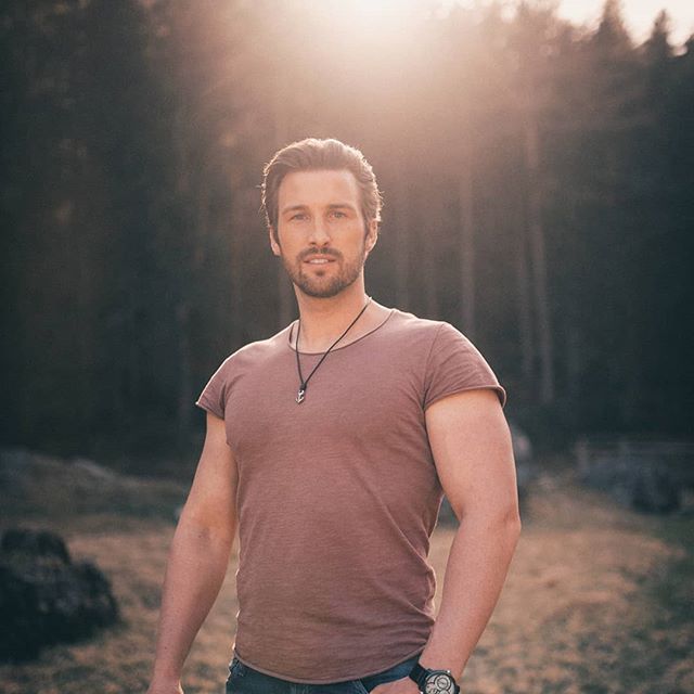 A confident man stands in a sunlit forest, blending fitness with nature's tranquility.