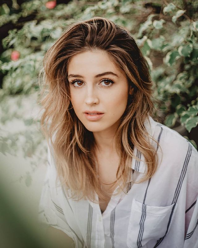 A young woman with brown hair poses in a garden, looking directly at the camera with a serene expression.