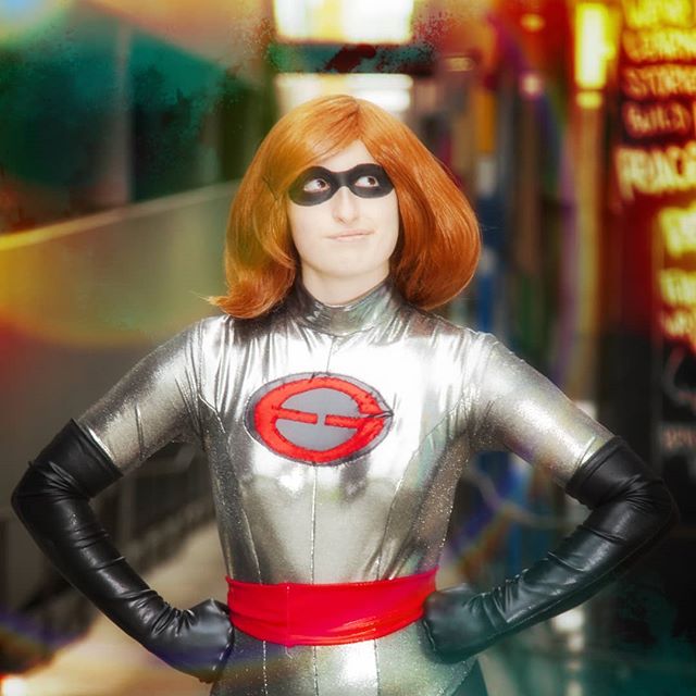 A woman dressed as a superhero with a silver suit, red belt, and black eye mask stands confidently in an urban setting.