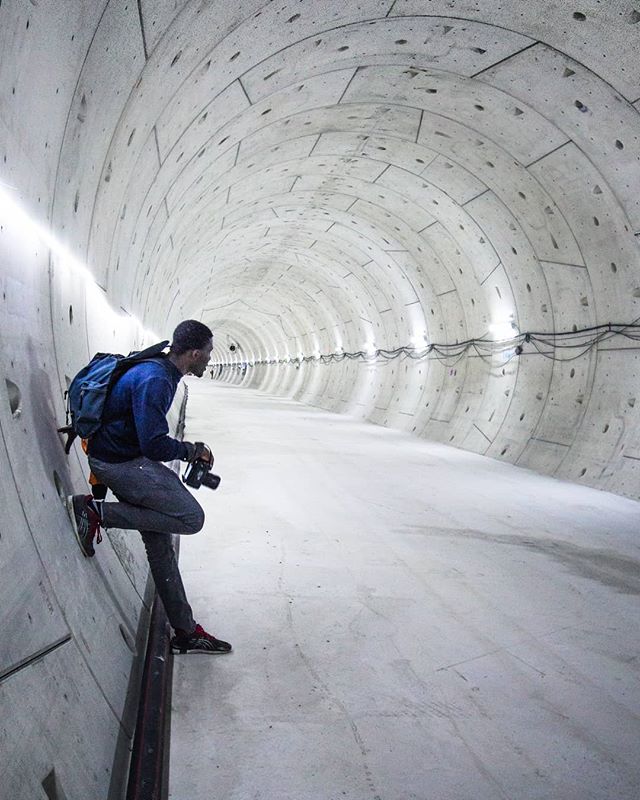 A man with a camera stands in a long, curved concrete tunnel, looking into the distance.