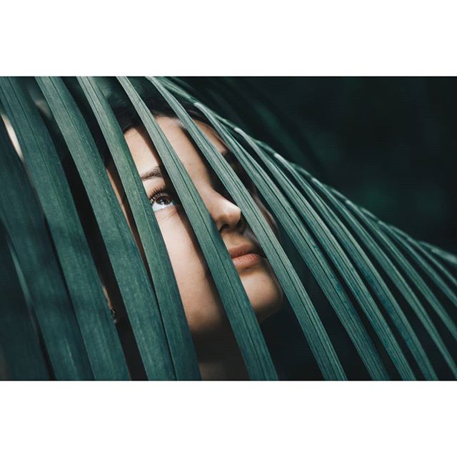 A woman peers through tropical green leaves, creating a dreamy, serene nature portrait.