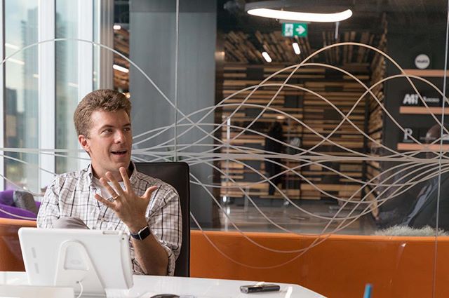 A man sits at a table in a modern office, using a tablet and gesturing during a discussion.