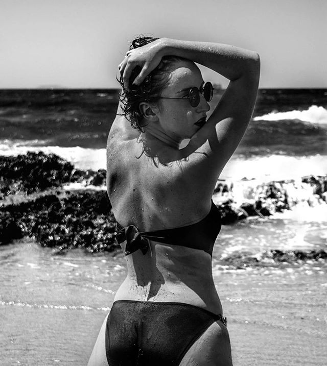 A woman in a bikini poses on a beach with ocean waves in the background in a black and white photograph.