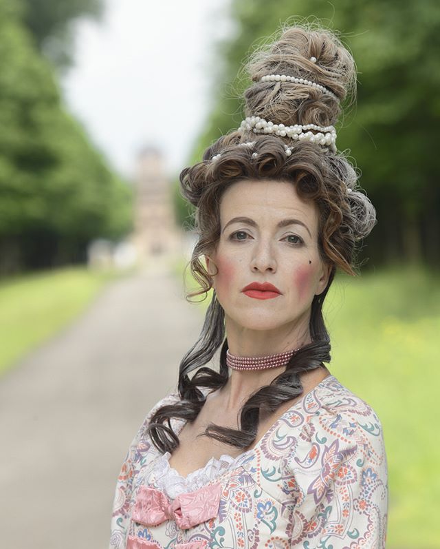 A woman in a rococo dress and hairstyle stands in a park. A building is visible in the distance.