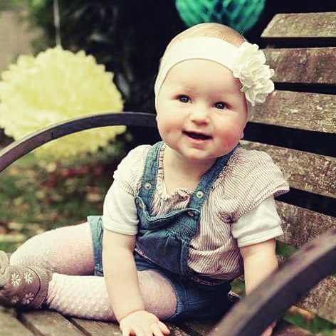 A happy baby with a headband sits on a park bench, radiating joy and warmth.