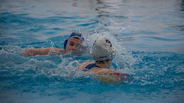 Two women compete in a water polo match in a bright blue pool.