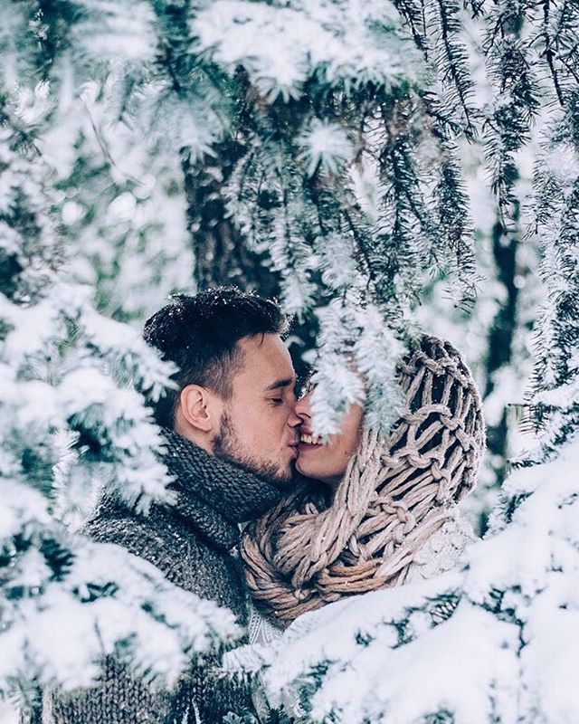 A couple shares a kiss in a snowy winter forest, surrounded by snow-covered trees in an intimate and romantic scene.