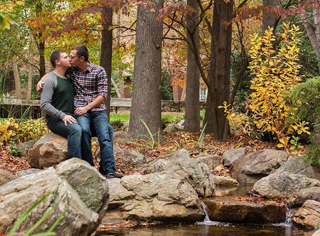A gay couple share a kiss sitting on rocks in a park surrounded by autumn leaves and trees.
