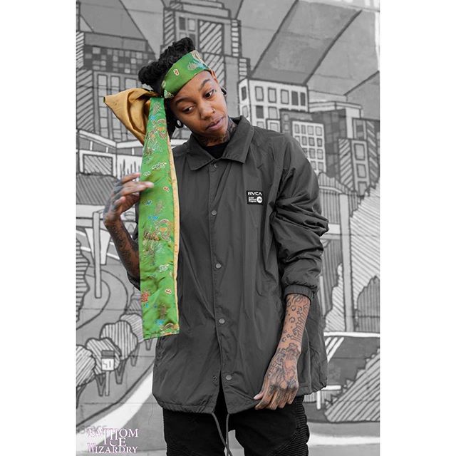 A tattooed person poses in a jacket and headscarf against an urban backdrop.