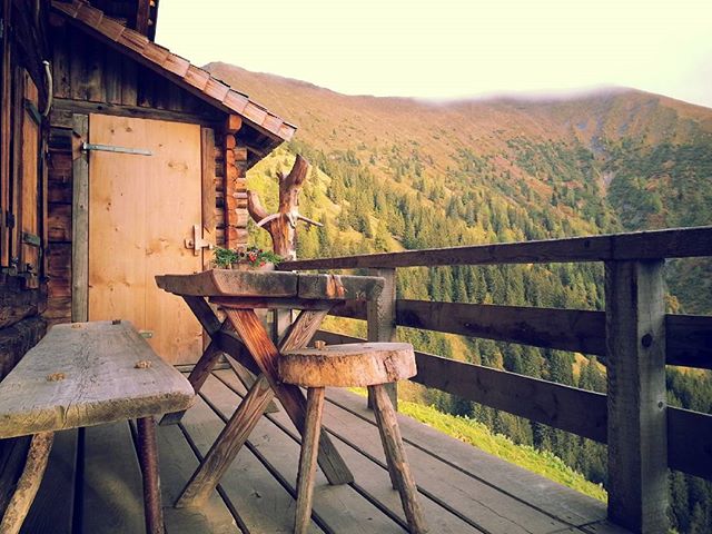 A rustic cabin deck offers a serene mountain view with a wooden table, bench and stool for a calming outdoor experience.
