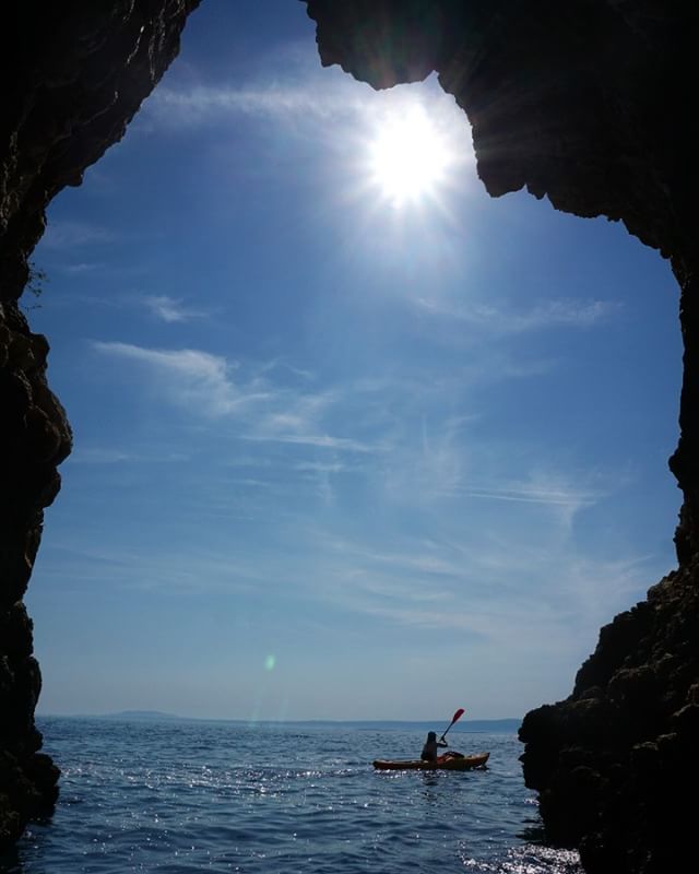 A person kayaking on the ocean, as seen through the opening of a cave with a bright sunny sky above.