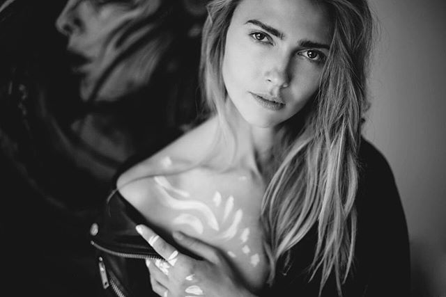 A black and white portrait of a blonde woman with artistic white paint on her skin, looking at the camera.