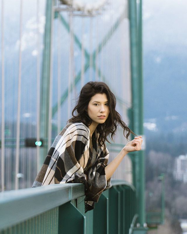 A brunette woman in a plaid shawl leans against a bridge railing with a cityscape and mountains in the background.