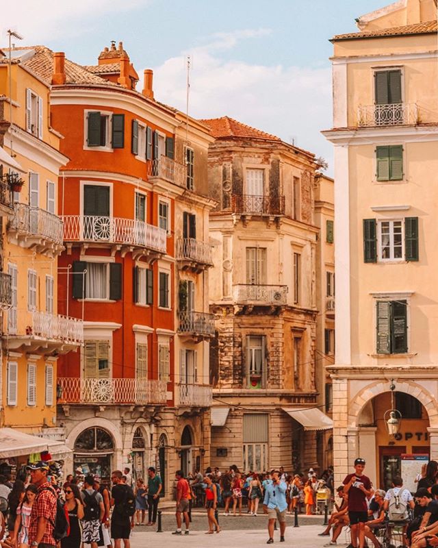A sunny European town square featuring colorful buildings and many people walking around enjoying the day.