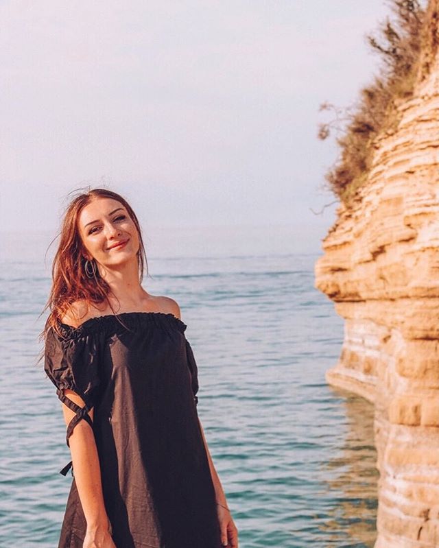 A woman in a black dress stands on a beach next to a rocky cliff overlooking the ocean.