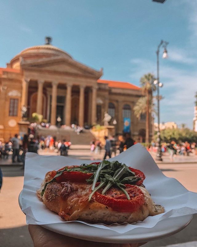A delicious pizza slice topped with tomatoes and arugula, with a historic building in the background.