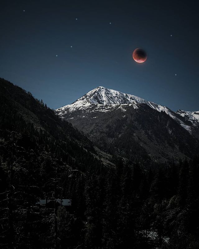 A blood moon rises above a snow-capped mountain peak on a clear night, creating a tranquil, mysterious scene.