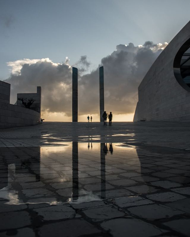 People walk through a modern plaza with monolithic structures reflected in puddles under a cloudy sky.