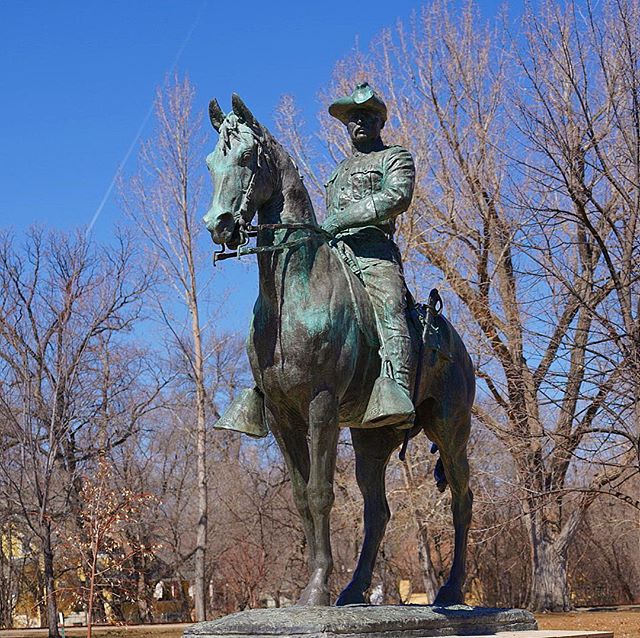 A bronze statue depicts a man on horseback against a clear blue sky and bare trees.