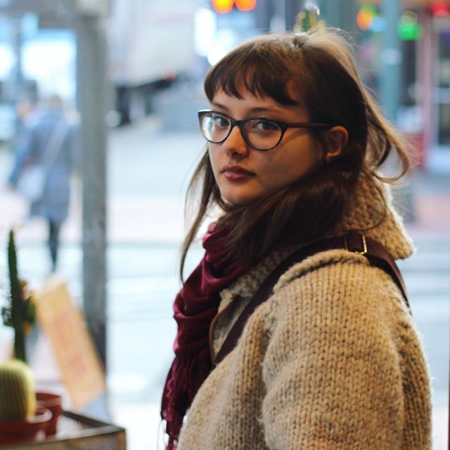 A woman wearing glasses and a scarf looks towards the camera in an urban setting. 