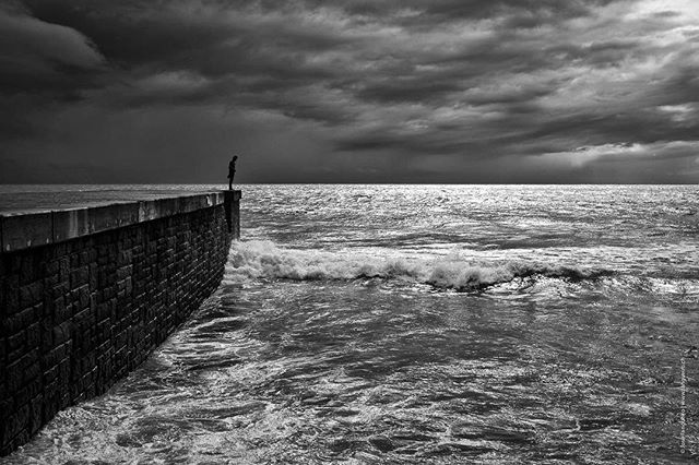 A lone man stands at the end of a pier overlooking the stormy sea under a dramatic sky.