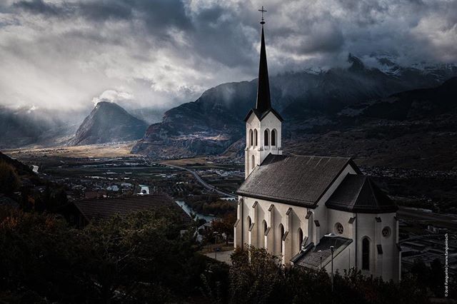 A scenic view of a church with a spire in a valley surrounded by mountains under a cloudy sky.