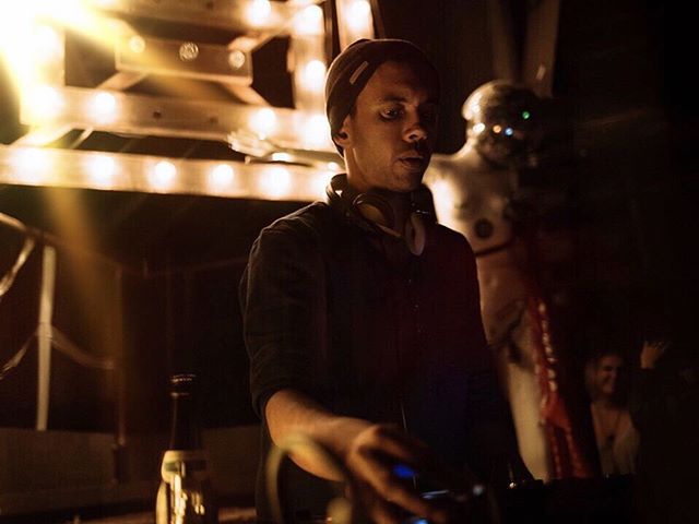 A DJ in a nightclub plays music with a light display in the background, creating an energetic party atmosphere.