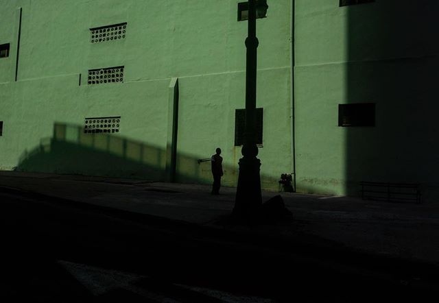 A solitary figure walks along a shadowed street with a green building, illuminated by a streetlamp.