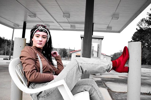 A stylish woman in jeans and a headscarf poses with a red boot at a vintage gas station.