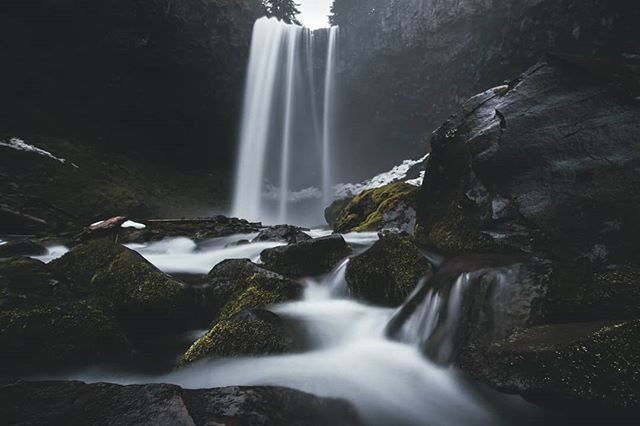 A scenic waterfall flows over moss-covered rocks in a lush forest, captured with a long exposure to emphasize the water's movement.