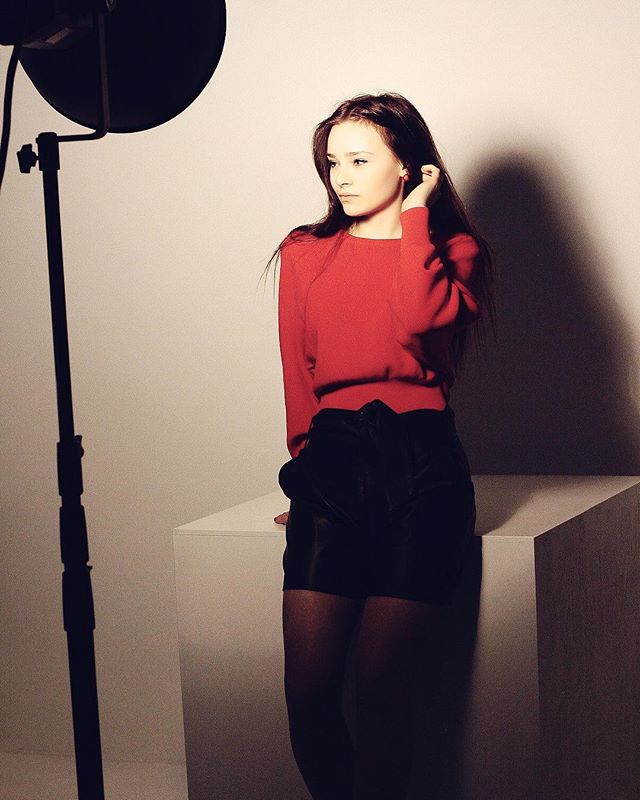 A young woman poses in a studio wearing a red sweater and black skirt.