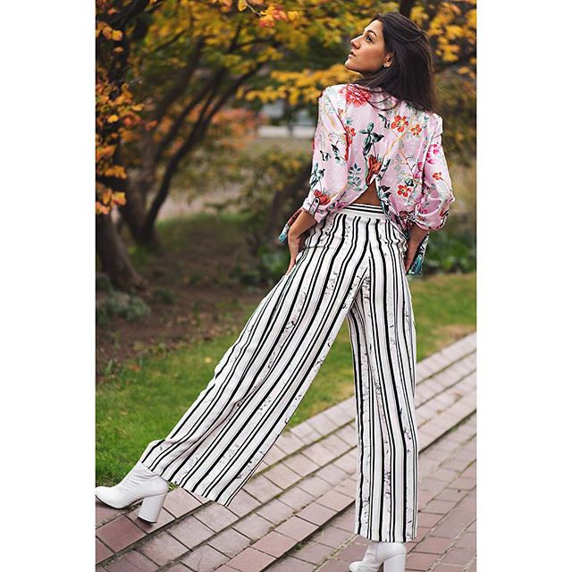 A woman in a floral blouse and striped pants poses in a park during autumn, showcasing stylish fall fashion.